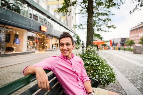 Preview: Smiling man sitting on bench by road against buildings in city