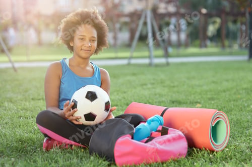 Preview: Cheerful little girl with soccer ball sitting on grass outdoors