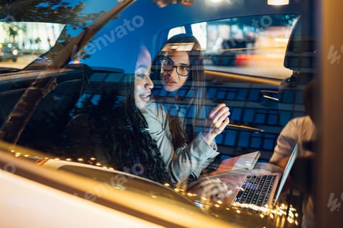Preview: Two multiracial business woman riding in a car and using a laptop