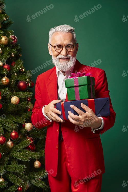 Preview: classy cheerful Santa in red suit posing next to pine tree holding pile of presents, winter concept