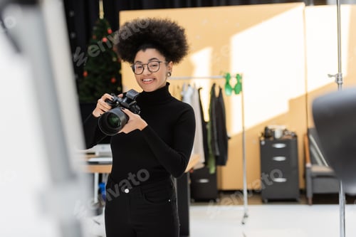 Preview: pleased african american photographer standing with professional digital camera in photo studio