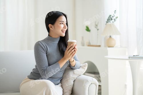 Preview: Young Asian woman drinking coffee while sitting at her couch.