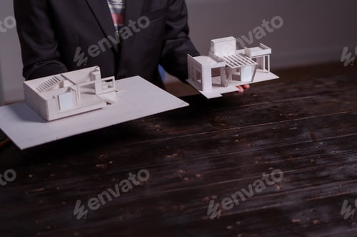 Preview: A businessman sits at a table at night, holding a model of a two-story white house. He reviews