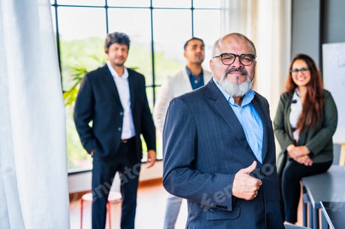 Preview: Confident oldIndian business leader standing with folded hands, colleagues out of focus in office