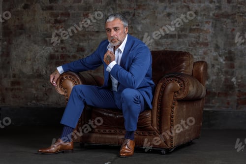 Preview: A man in a blue formal suit sitting on a leather armchair.Portrait of a businessman on a brick wall