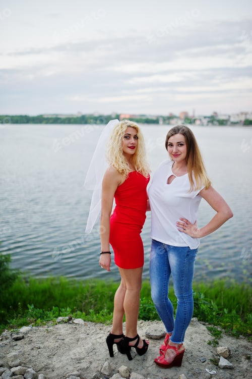 Preview: Awesome bride in red dress posing with her bridesmaid on the lakeside.