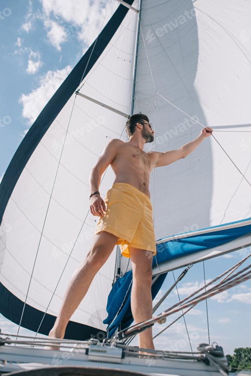 Preview: low angle view of shirtless muscular man in swim trunks adjusting sail on yacht