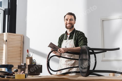 Preview: Smiling restorer in apron holding sandpaper near chair and looking at camera in workshop