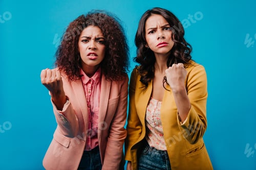 Preview: Indoor portrait of two girls waving their fists. Studio shot of angry women isolated on blue backgr