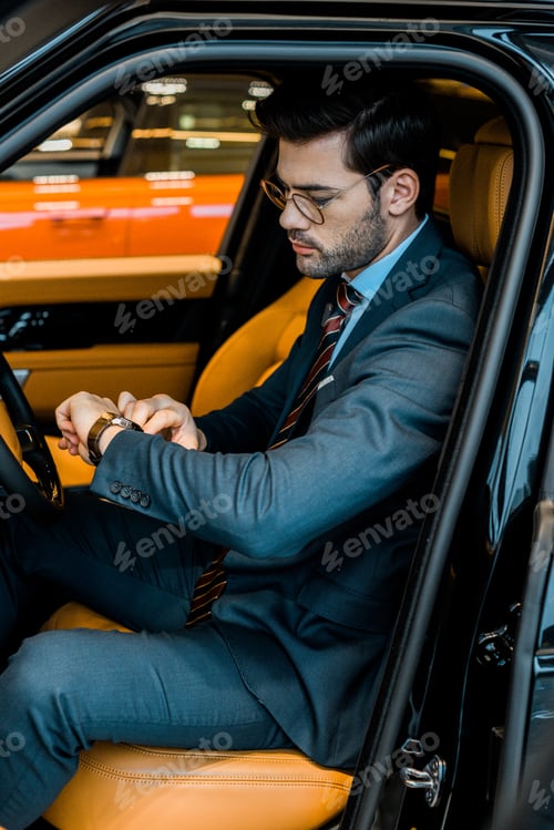 Preview: handsome businessman in eyeglasses checking watch in luxury car