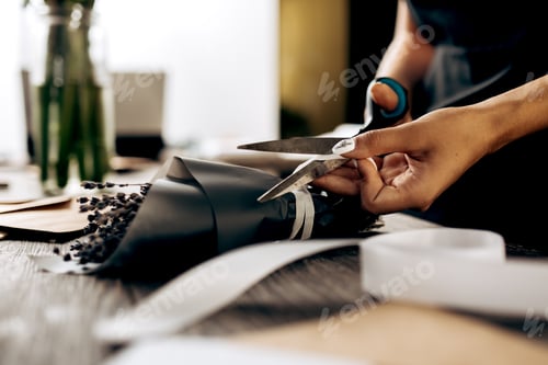 Preview: Florist cuts a ribbon on the little bouquet of flowers packed in dark paper on the wooden table with