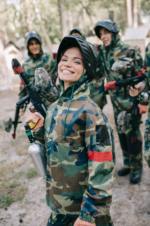 Preview: attractive smiling female paintballer in uniform holding paintball gun while her team standing