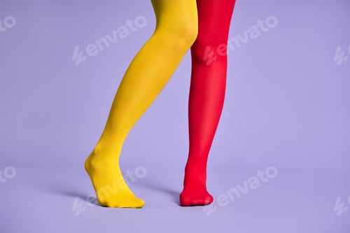 Preview: Long slim woman legs in multicolor yellow and red tights against purple background.