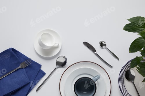 Preview: top view of green leaves, various ceramic plates, cup, kitchen towel, fork, spoons and knife on