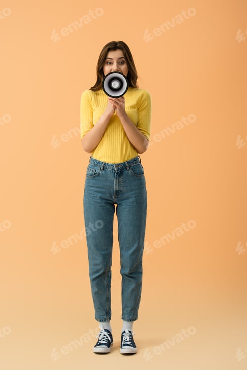 Preview: Full length view of brunette girl in jeans screaming in megaphone on orange background