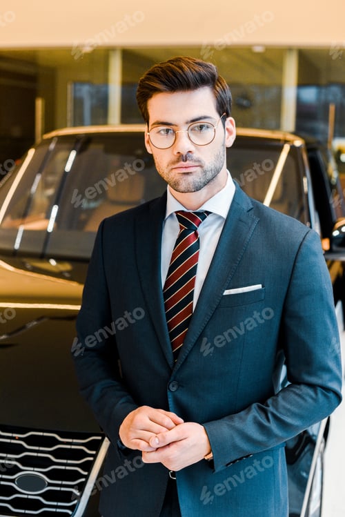 Preview: selective focus of young businessman in eyeglasses looking at camera near car in showroom