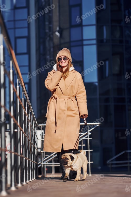 Preview: Woman in warm clothes have a walk with her little pug dog near business building that on background