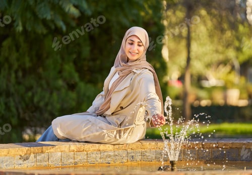 Preview: an Arab woman with a headscarf sitting on the edge of a fountain, touching the water with her hand