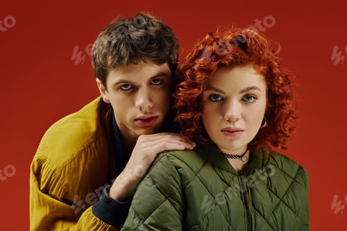 Preview: stylish loving couple in fashionable attires posing together and looking at camera on red backdrop