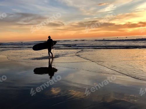 Preview: A surfer in a hat leaves the beach with his long surfboard at sunset