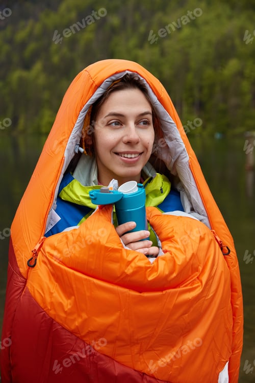 Preview: Lovely happy woman covered with warm sleeping bag, drinks hot tea or coffee, focused into distance w