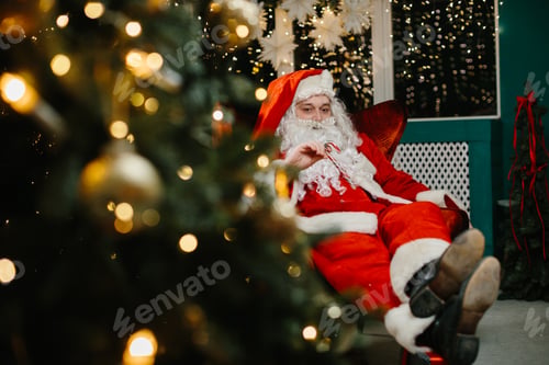 Preview: Santa claus relaxing in armchair with candy cane near christmas tree