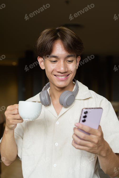 Preview: Non binary Asian student man sitting in cafe drinking coffee and using phone
