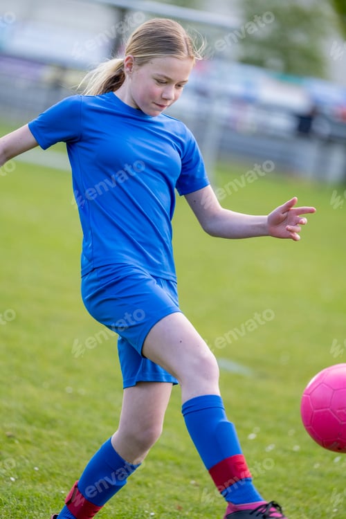 Preview: Young female soccer player in blue uniform kicking a pink ball on a grassy field.