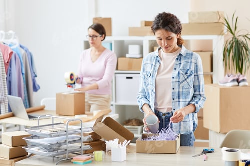 Preview: Pretty young manager of online shop packing mug in carton box