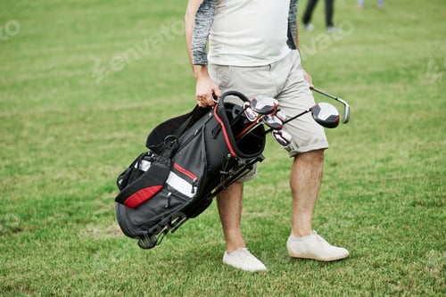 Preview: Cropped photo of man holding bag with golf sticks while walking on the green lawn