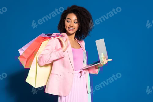 Preview: smiling elegant african american businesswoman with shopping bags using laptop on blue background