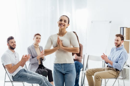 Preview: smiling woman looking at camera while people sitting on chairs during group therapy session