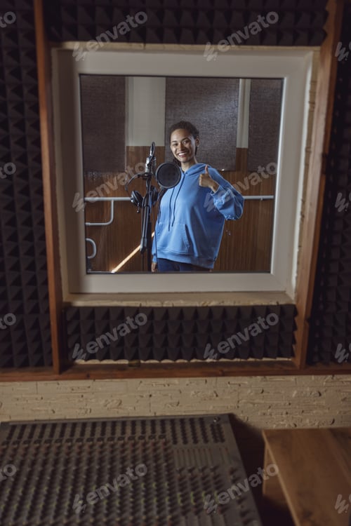 Preview: Woman singer shows thumb up standing near microphone inside a soundproof room of recording studio