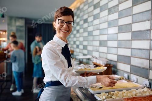 Preview: Happy waitress serving food during breakfast at hotel restaurant and looking at camera.