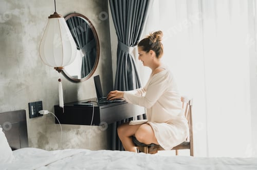 Preview: Full body side view of a young woman in a white robe sitting on a chair at a table while browsing th