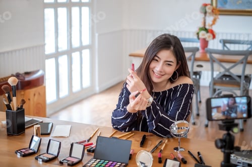 Preview: Young asian woman blogger with makeup cosmetics recording video clip online by smartphone at home.