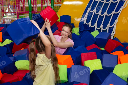 Preview: Happy millennial mother and her teen daughter having fight with foam cubes at indoor kids