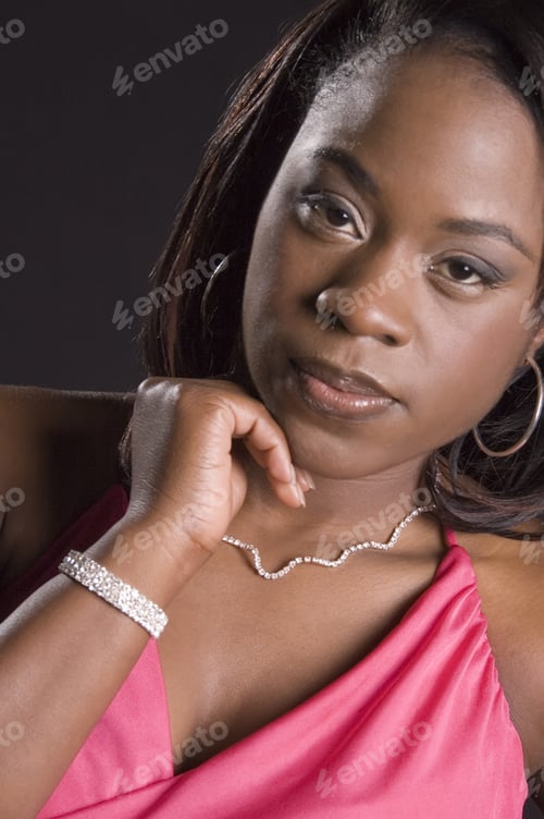 Preview: Young Black Woman In Pink Dress And Jewelry Resting Chin On Fingers