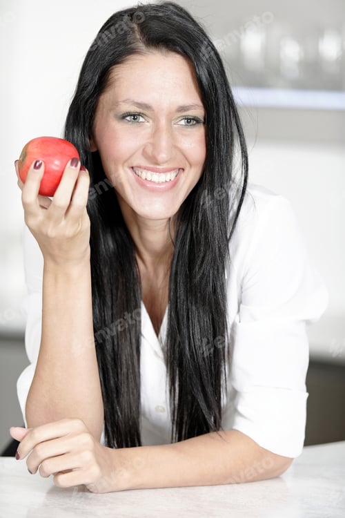 Preview: Beautiful Young Woman Eating An Apple In Her Kitchen At Home