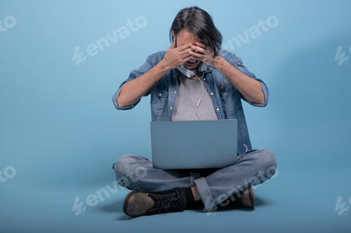 Preview: Young Bearded Man Sit Down On Floor Using Computer In Blue Background. Upset Asian Young Hipster
