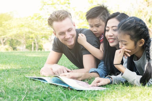 Preview: Family Reading A Book Together In Park. Mixed Race Family Laying Down Together Reading Book. White