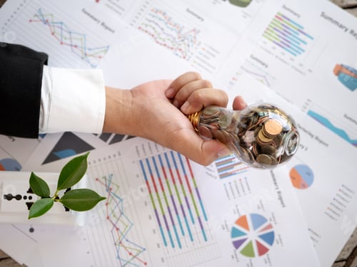 Preview: Top View Hand Of Businessman Hold Light Bulb With Coin And Calculator On Business Graph. Concept Of