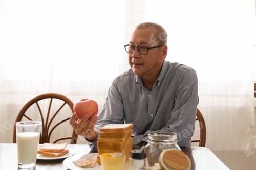 Preview: Asian Elderly Man Having Bread Breakfast, Milk And Fruit With Carefully In The Kitchen Room At Home