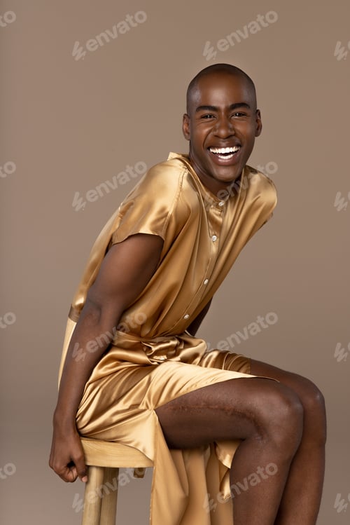 Preview: Handsome Rwandese Man In His 20S Wearing A Gold Tunic Posed Sitting On A Stool On A Neutral