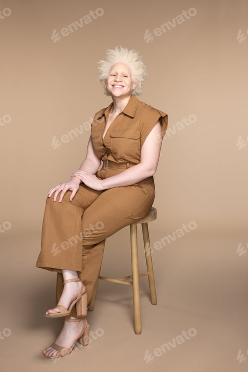 Preview: Studio Shot Of Beautiful Black Non-Binary Person With Albinism In Their 20S Sitting On Stool And