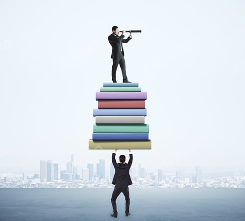 Preview: Businessman Holding Book Stack With Colleague Looking Into The Distance With Telescope On Blurry