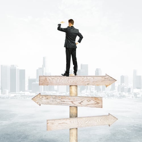Preview: Businessman Standing On Abstract Wooden Road Sign. City Background. Different Direction Concept