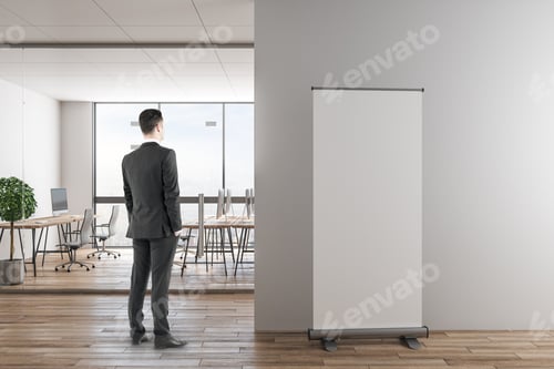 Preview: Young Businessman Standing In Modern Office Interior With Empty Banner On Wall. Mock Up,