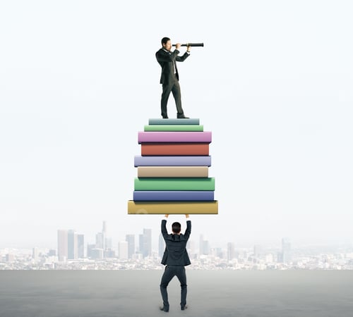 Preview: Businessman Holding Book Stack With Colleague Looking Into The Distance With Telescope On Blurry