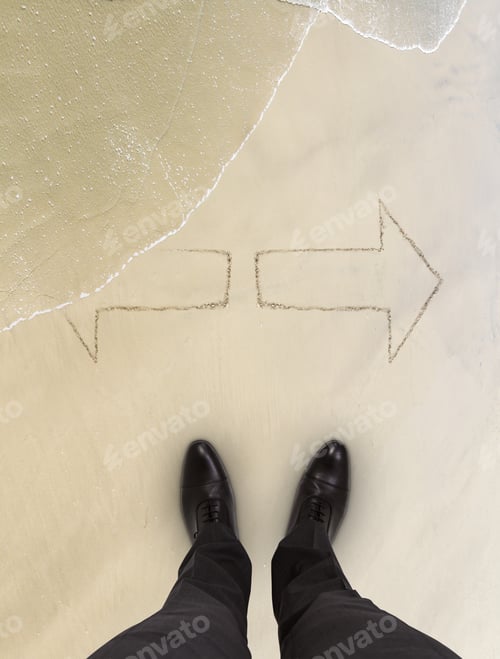 Preview: Businessman Standing On Beach And Looks At Arrows On Sand